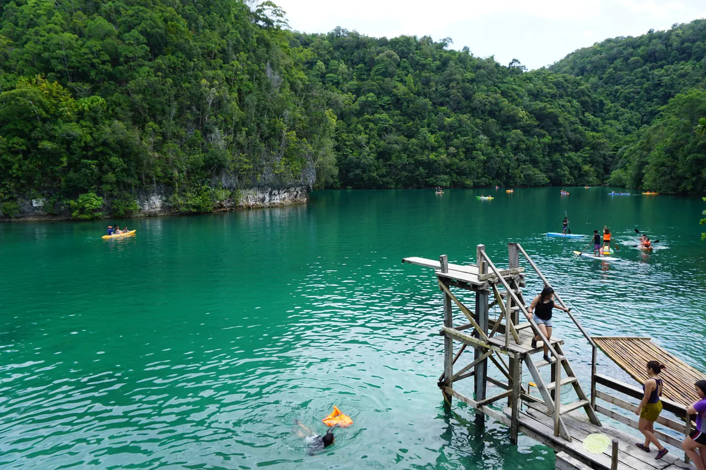 Photo: Lucky Ambago Purok Otso (CC BY-SA 4.0) Green water of Sugba Lagoon in Del Carmen, ringed by forested limestone hills, with a wooden jumping platform, kayaks and paddleboards