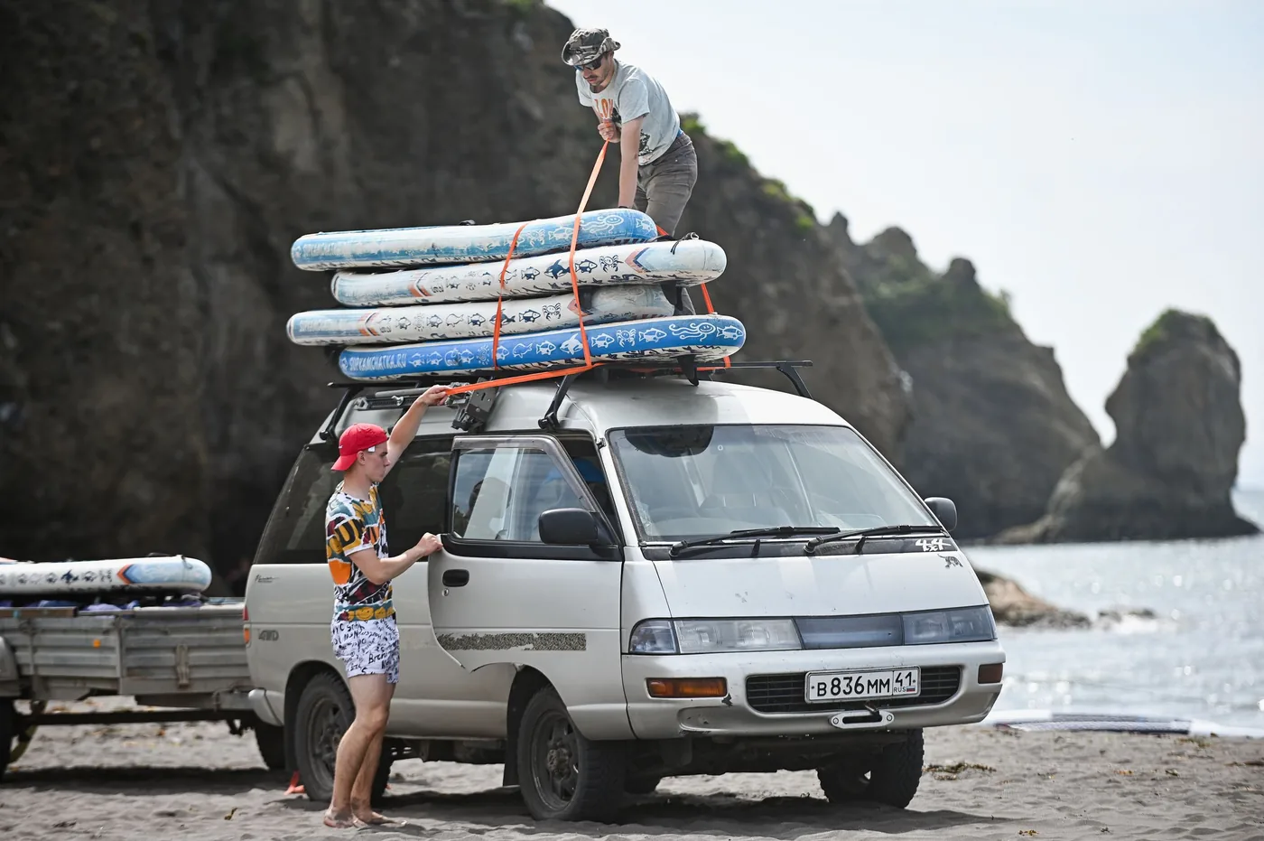 Two people lifting surfboards onto the roof rack of a van parked beside a beach